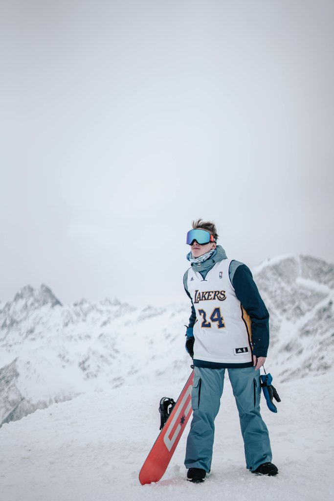 Snowboarder wearing Lakers jersey stands atop a snowy mountain, ready for adventure.